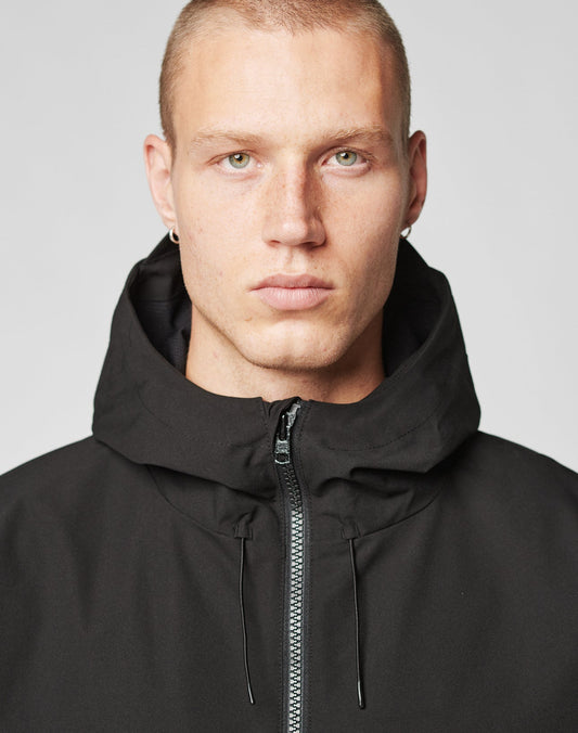A person with short blonde hair and light eyes wears the LIVE FAST DIE YOUNG Basic Shell Jacket in black, oversize fit, zipped to the neck, staring at the camera against a plain light background.