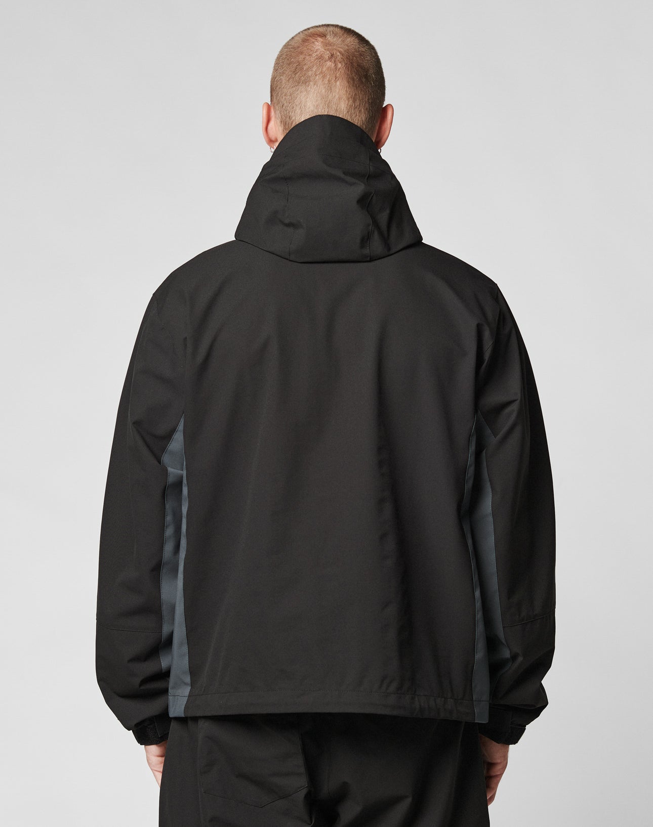 A person wearing the LIVE FAST DIE YOUNG Basic Shell Jacket, featuring an oversize fit, black color, dark gray side panels, and a water-repellent zipper, is shown from the back against a plain light background.