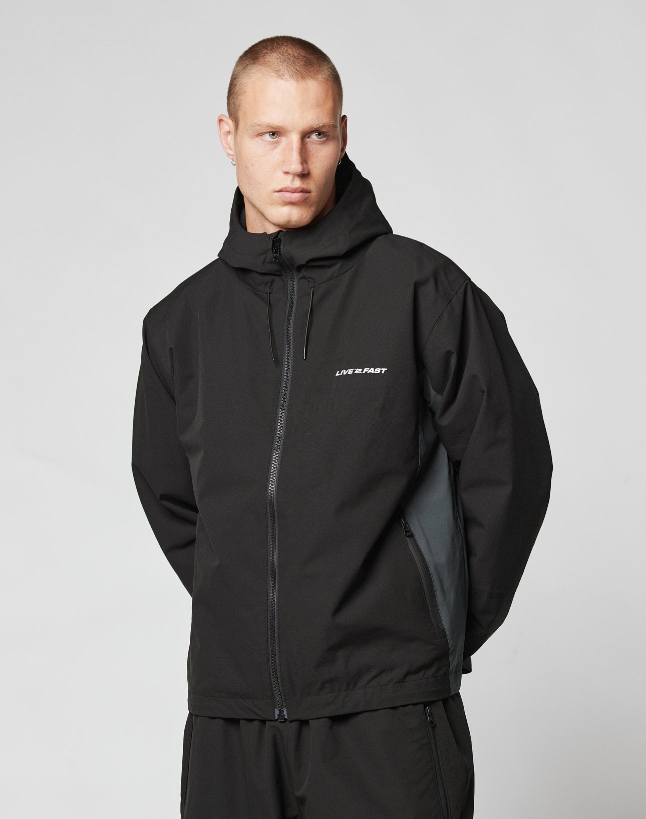 A person with short hair wears the LIVE FAST DIE YOUNG Basic Shell Jacket in black, featuring reflektierender Druck and LIVE FAST in white on the chest, standing against a plain light background with hands behind their back.