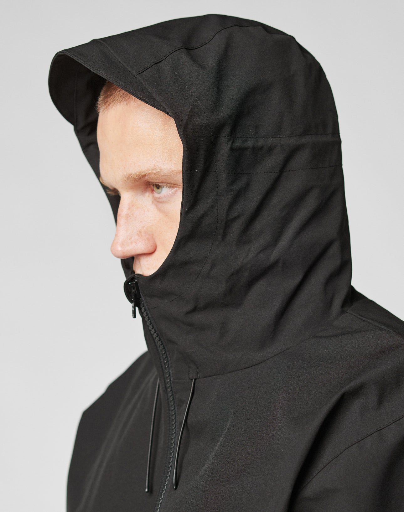 A person wearing the LIVE FAST DIE YOUNG Basic Shell Jacket, black with hood up, shown from the side against a plain background. The oversize fit and water-resistant zipper offer comfort and protection, partially covering the face.