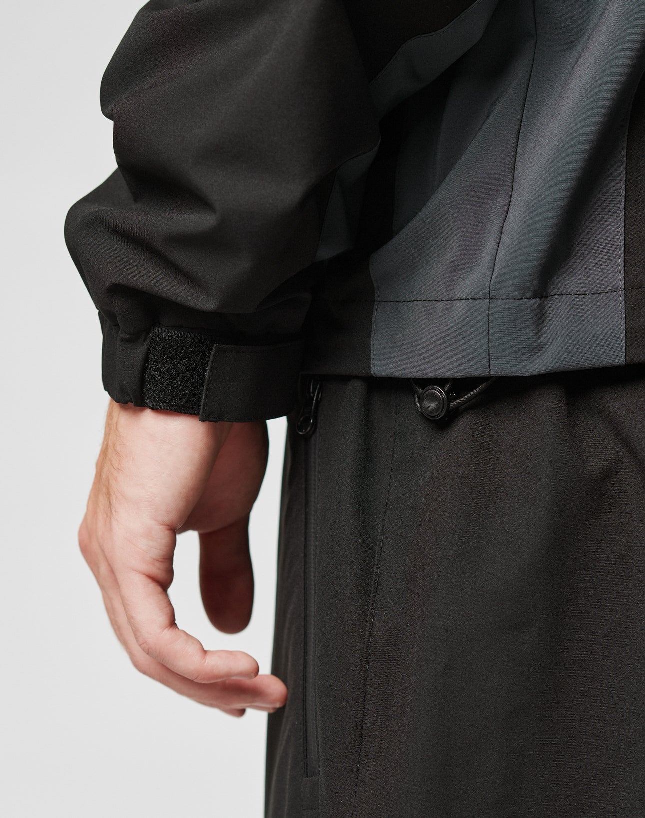 Close-up of a left hand and wrist wearing the LIVE FAST DIE YOUNG Basic Shell Jacket with a wasserabweisendem Reißverschluss, paired with matching dark pants. The light background highlights its practical details.