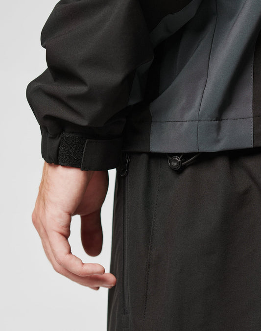 Close-up of a left hand and wrist wearing the LIVE FAST DIE YOUNG Basic Shell Jacket with a wasserabweisendem Reißverschluss, paired with matching dark pants. The light background highlights its practical details.
