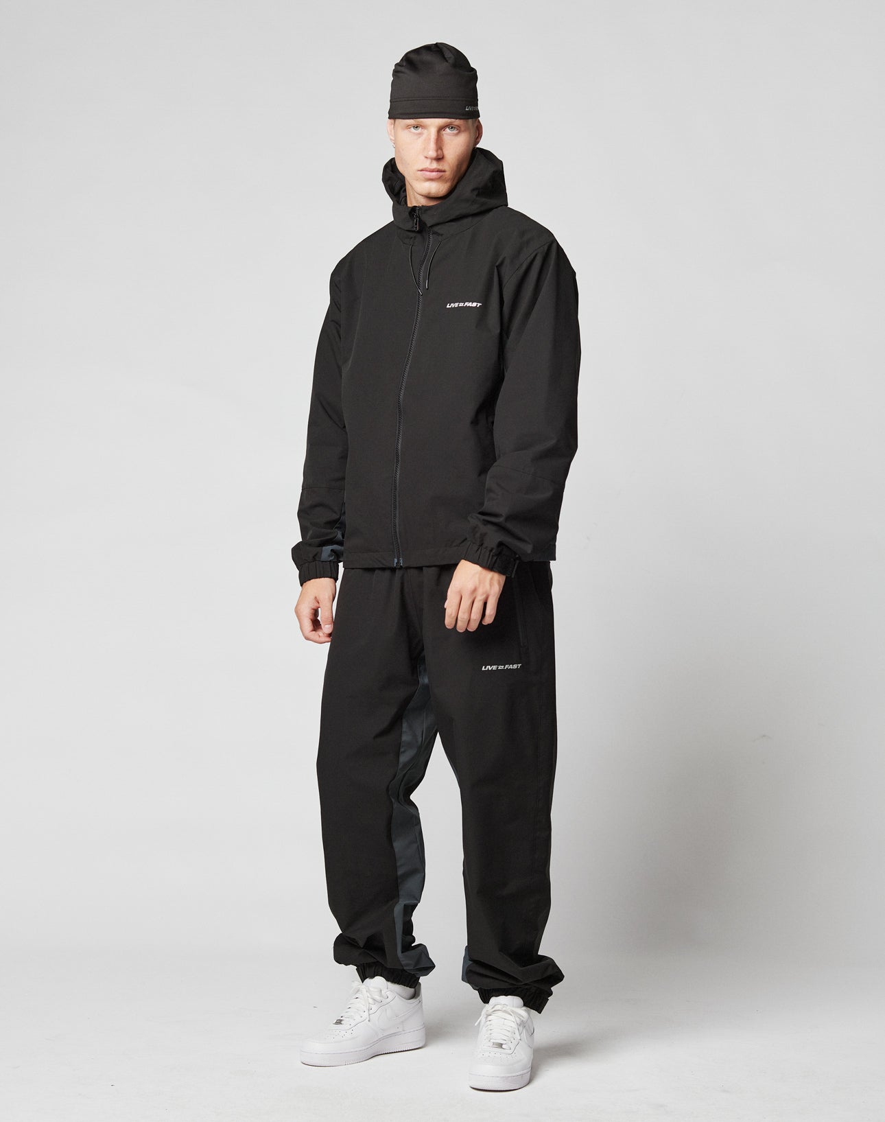 A person wears LIVE FAST DIE YOUNG Basic Shell Pants with an elastic waistband and drawstring, a black oversize fit jacket, black beanie, and white sneakers against a light gray background for a sporty, comfortable look.