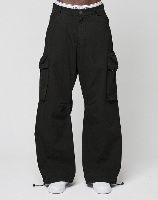 A person wears the LFDY Baggy Cargo by LIVE FAST DIE YOUNG—loose black cargo pants with großen Cargo-Taschen, a glimpse of a LIVE FAST waistband, and white sneakers—posing against a plain white background. 100% Baumwolle für Komfort.