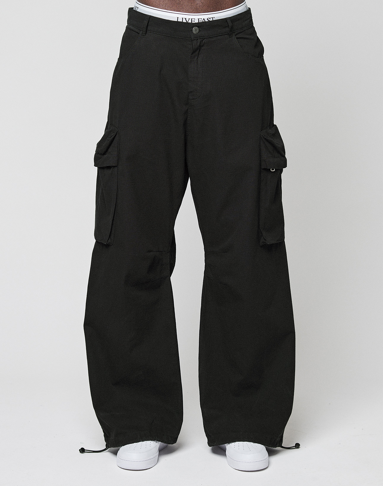A person wears the LFDY Baggy Cargo by LIVE FAST DIE YOUNG—loose black cargo pants with großen Cargo-Taschen, a glimpse of a LIVE FAST waistband, and white sneakers—posing against a plain white background. 100% Baumwolle für Komfort.