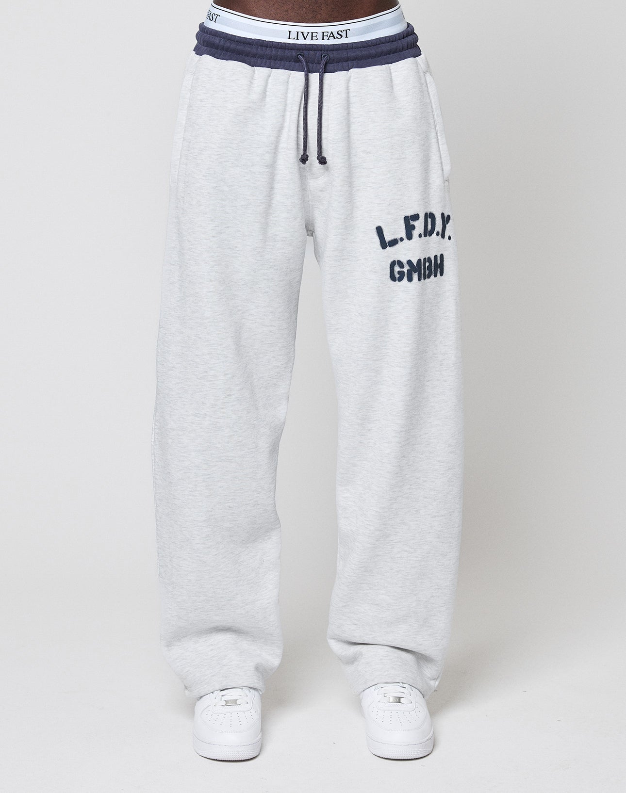 A person wears the LFDY District Jogger by LIVE FAST DIE YOUNG in light gray (wide fit) with dark blue "L.F.D.Y. GMBH" on the left thigh, zippered side pockets, white sneakers, and a visible LIVE FAST waistband.