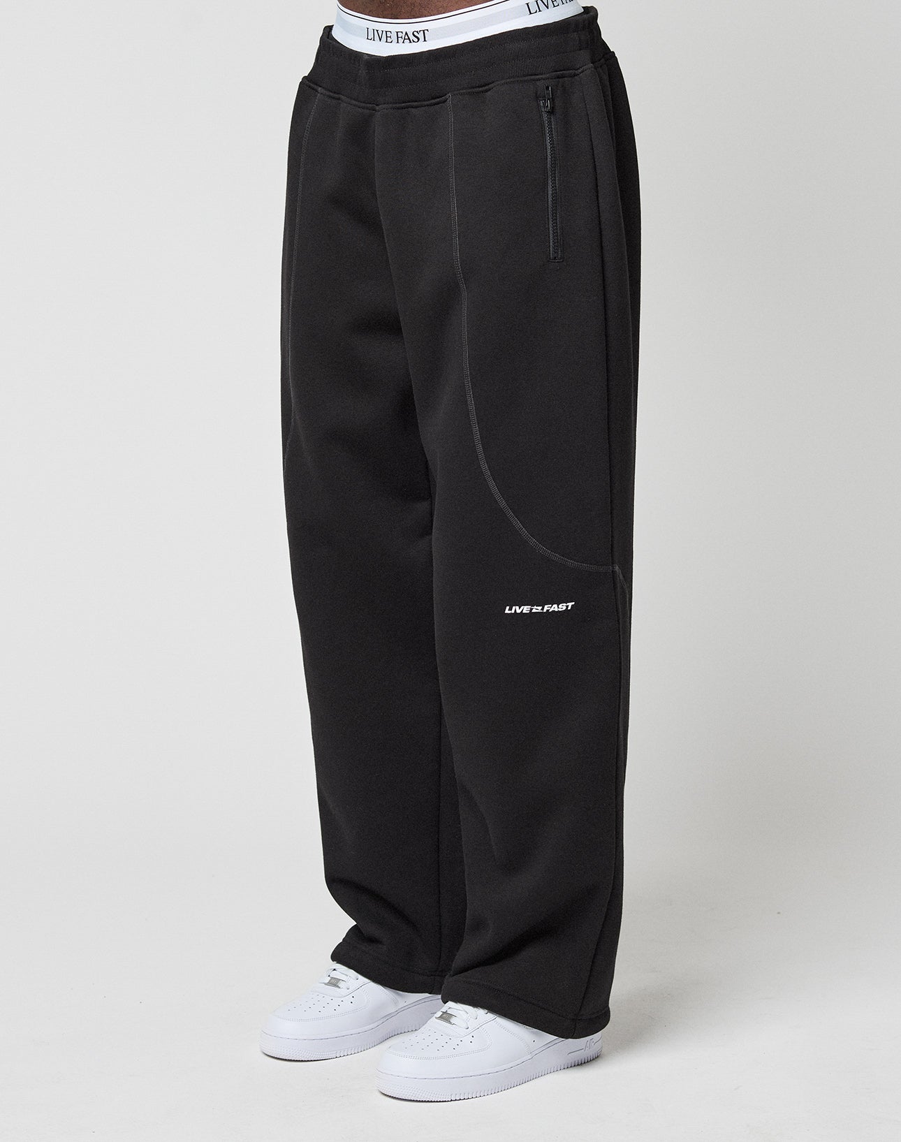 Model wears the LFDY Active Fleece Jogger by LIVE FAST DIE YOUNG—wide fit, black with LIVE FAST logo and branded elastic waistband—paired with white sneakers, standing against a plain white background. | model