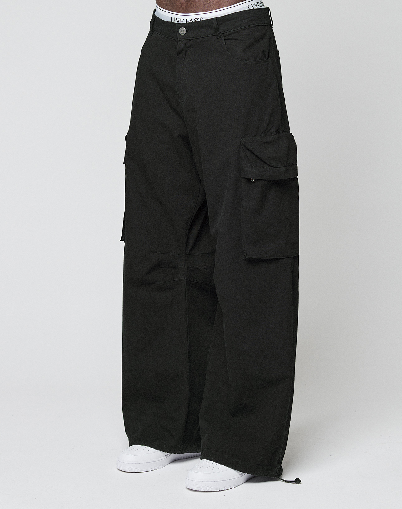 Eine Person trägt die LFDY Baggy Cargo von LIVE FAST DIE YOUNG aus 100% Baumwolle mit großen Cargo-Taschen, sichtbarem weißem Bund und weißen Sneakers vor einem schlichten, hellen Hintergrund. | model