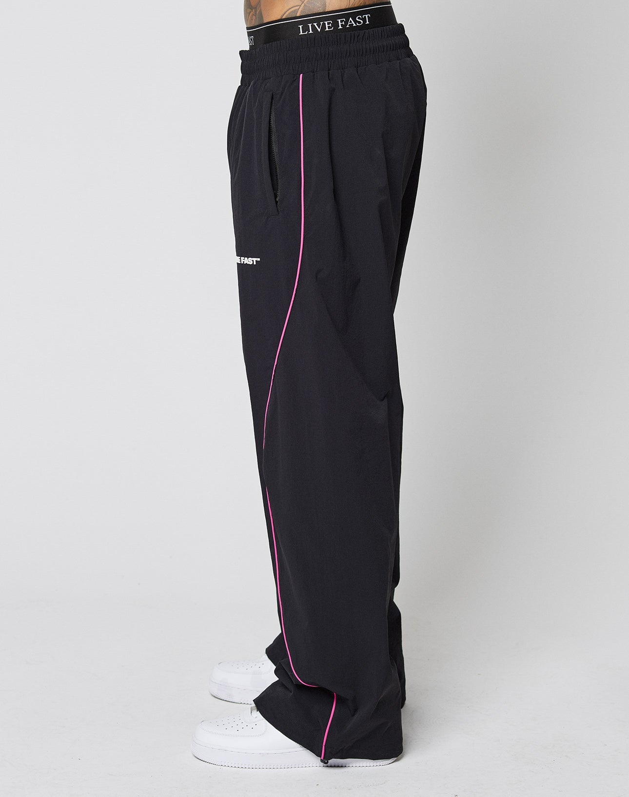A person stands sideways in LFDY Wavy Trackpants by LIVE FAST DIE YOUNG—wide fit black pants with pink piping and a LIVE FAST logo on the elastic waistband—paired with white sneakers, against a plain white background.