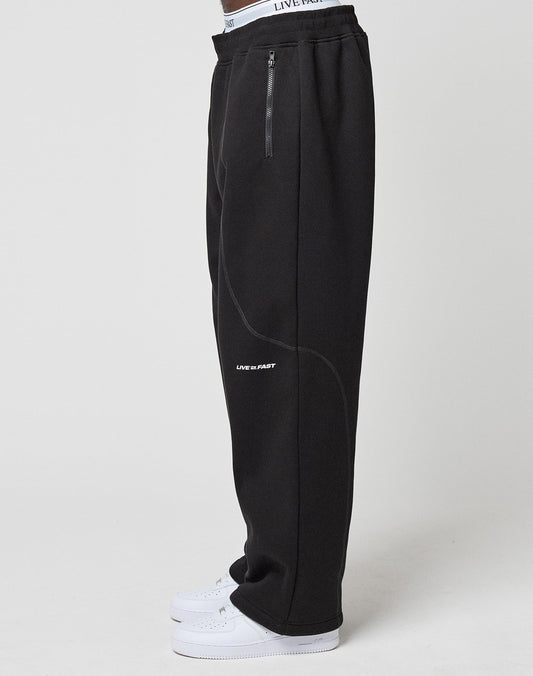 A person, shown from the waist down, models the LFDY Active Fleece Jogger by LIVE FAST DIE YOUNG—wide fit black sweatpants with zip pockets and a bold LIVE FAST logo on the thigh—paired with white sneakers against a plain background.