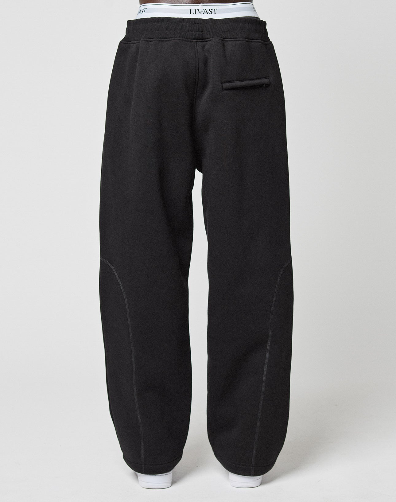 Back view of a person wearing the LFDY Active Fleece Jogger by LIVE FAST DIE YOUNG in wide fit black with contrast stitching, a single back pocket, and visible white waistband. The plain background highlights its relaxed style.