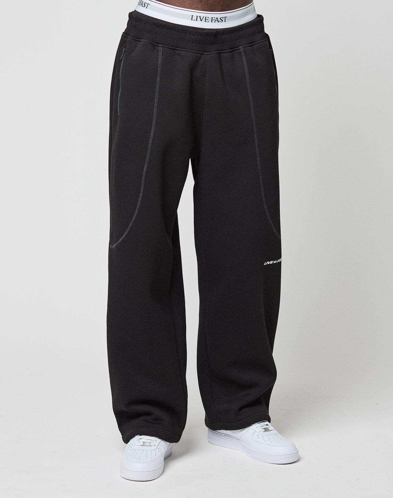 A person wears loose black LFDY Active Fleece Joggers by LIVE FAST DIE YOUNG, featuring subtle seam details and a Wide Fit. The white LIVE FAST waistband peeks above the pants, paired with white sneakers, against a plain light background.