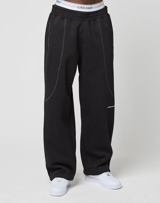 A person wears loose black LFDY Active Fleece Joggers by LIVE FAST DIE YOUNG, featuring subtle seam details and a Wide Fit. The white LIVE FAST waistband peeks above the pants, paired with white sneakers, against a plain light background.