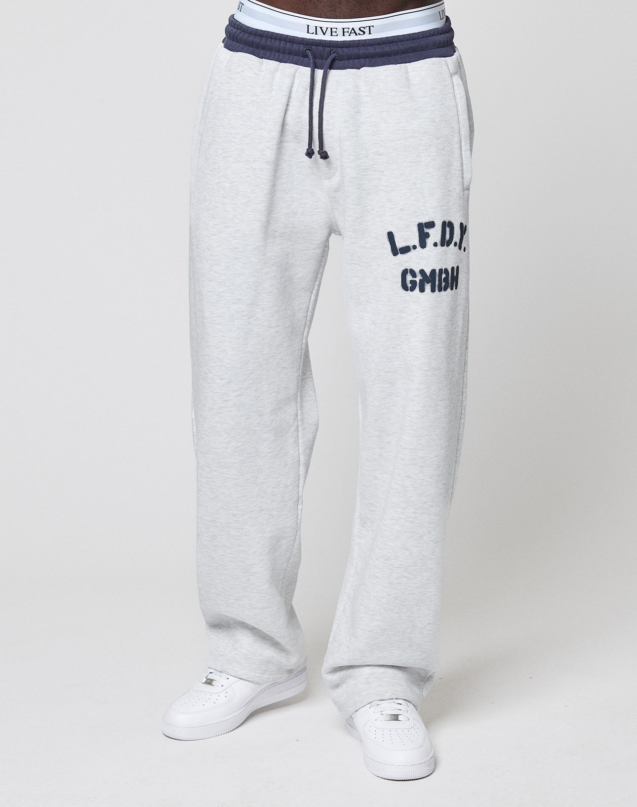 Someone is wearing LFDY District Jogger sweatpants by LIVE FAST DIE YOUNG in light gray with navy L.F.D.Y. GmbH logo on the left thigh, side zip pockets, white sneakers, and LIVE FAST underwear waistband visible above the pants.