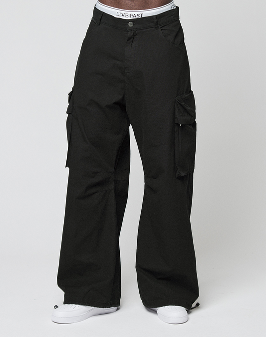 A person wears black LFDY Baggy Cargo pants by LIVE FAST DIE YOUNG with a baggy fit, white sneakers, and a visible LIVE FAST waistband, standing against a plain white background.