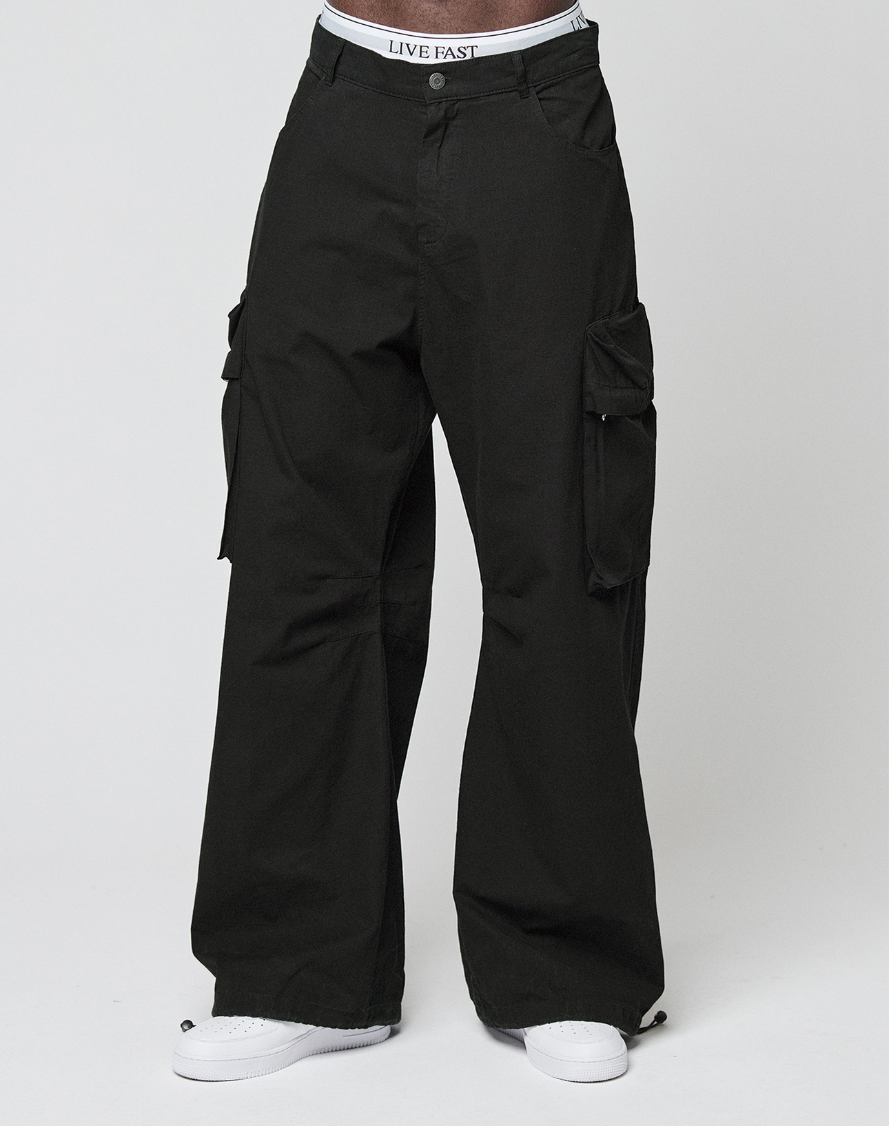 A person wears black LFDY Baggy Cargo pants by LIVE FAST DIE YOUNG with a baggy fit, white sneakers, and a visible LIVE FAST waistband, standing against a plain white background.