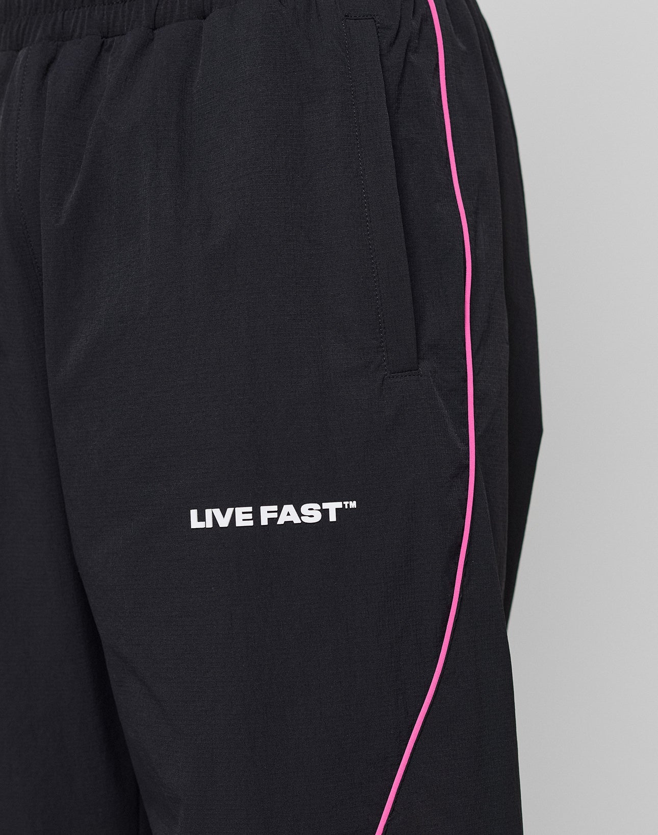 Close-up of LIVE FAST DIE YOUNGs LFDY Wavy Trackpants: black athletic pants with a white LIVE FAST™ logo, thin pink side stripe, elastic waistband, wide fit, and side pocket.