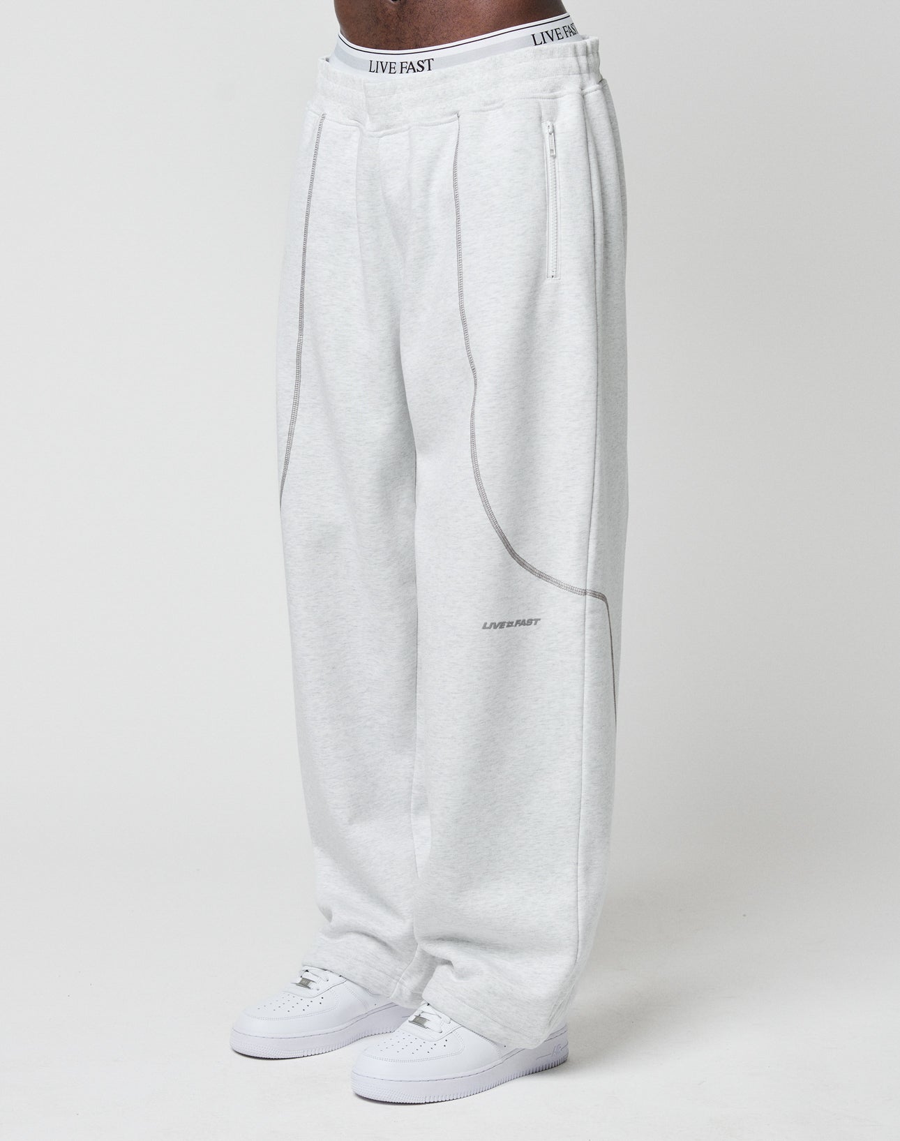 A person wears LFDY Active Fleece Jogger by LIVE FAST DIE YOUNG, featuring a wide fit, light gray color, curved seam details, zip pockets, logo print, and LIVE FAST text on the waistband and thigh, paired with white sneakers against a white background. | model