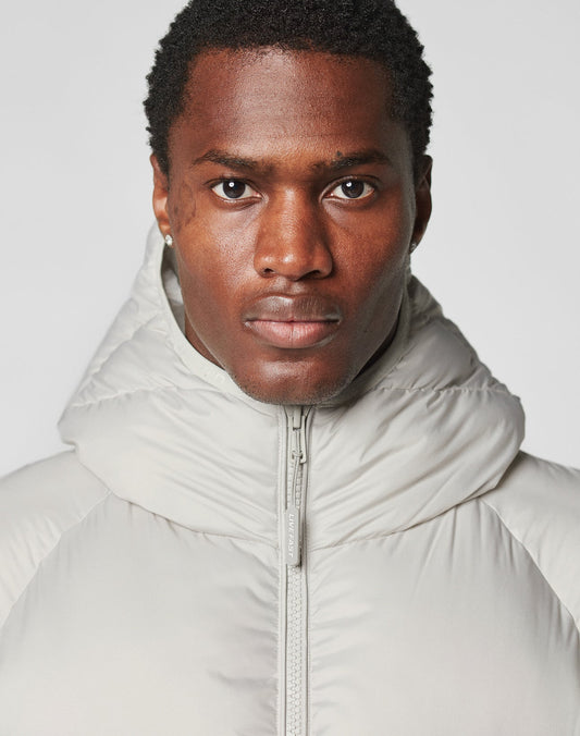 A person with short, curly hair wears the LIVE FAST DIE YOUNG Live Fast Puffer Jacket in light gray, featuring a hood-up and oversize fit, looking directly at the camera against a plain background.