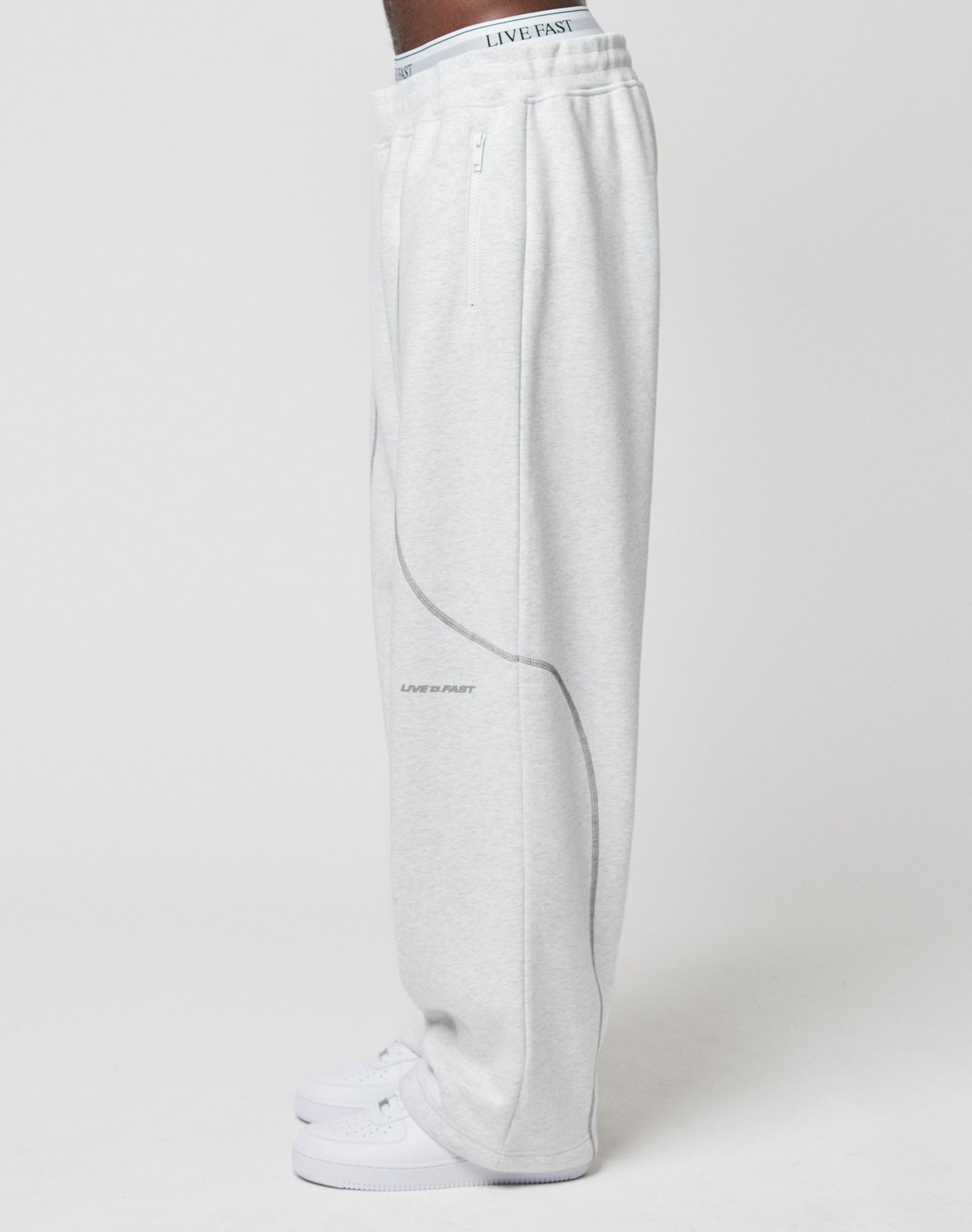 A person wearing LFDY Active Fleece Jogger by LIVE FAST DIE YOUNG—light gray with Logo Print LIVE FAST on waistband and leg, plus Reißverschlusstaschen—pairs them with white sneakers, standing in profile against a plain white background.