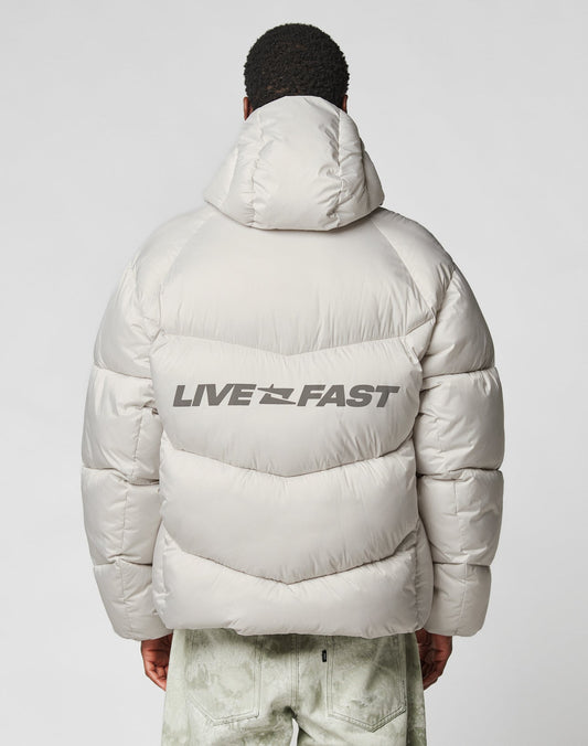 A person seen from behind wears the LIVE FAST DIE YOUNG Live Fast Puffer Jacket in light gray with an oversize fit, hood, and bold black reflective LIVE FAST print on the back. Light-colored pants finish the outfit.