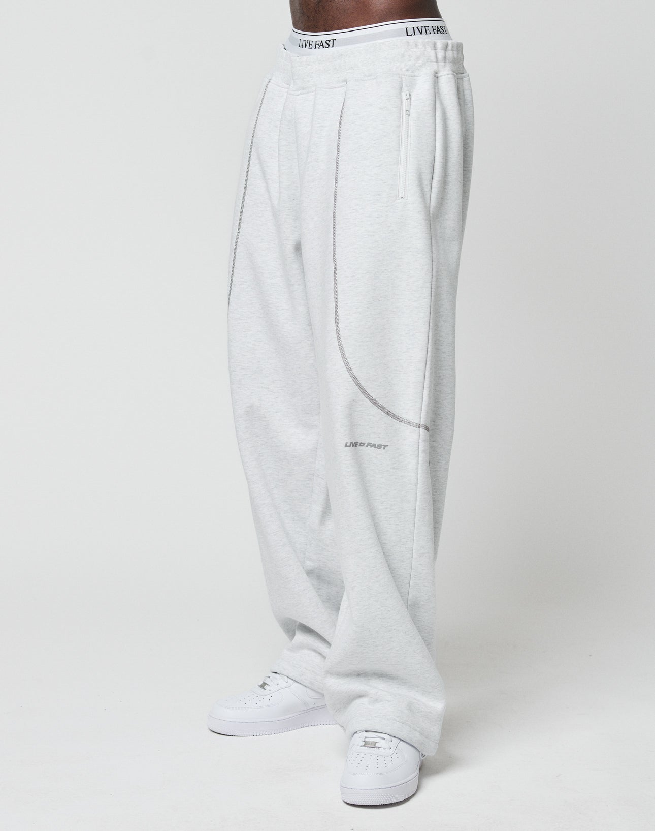 Person wears LFDY Active Fleece Jogger by LIVE FAST DIE YOUNG in wide fit light gray with subtle seams and small logo, paired with white sneakers. Visible: white waistband with LIVE FAST text. Plain white background.
