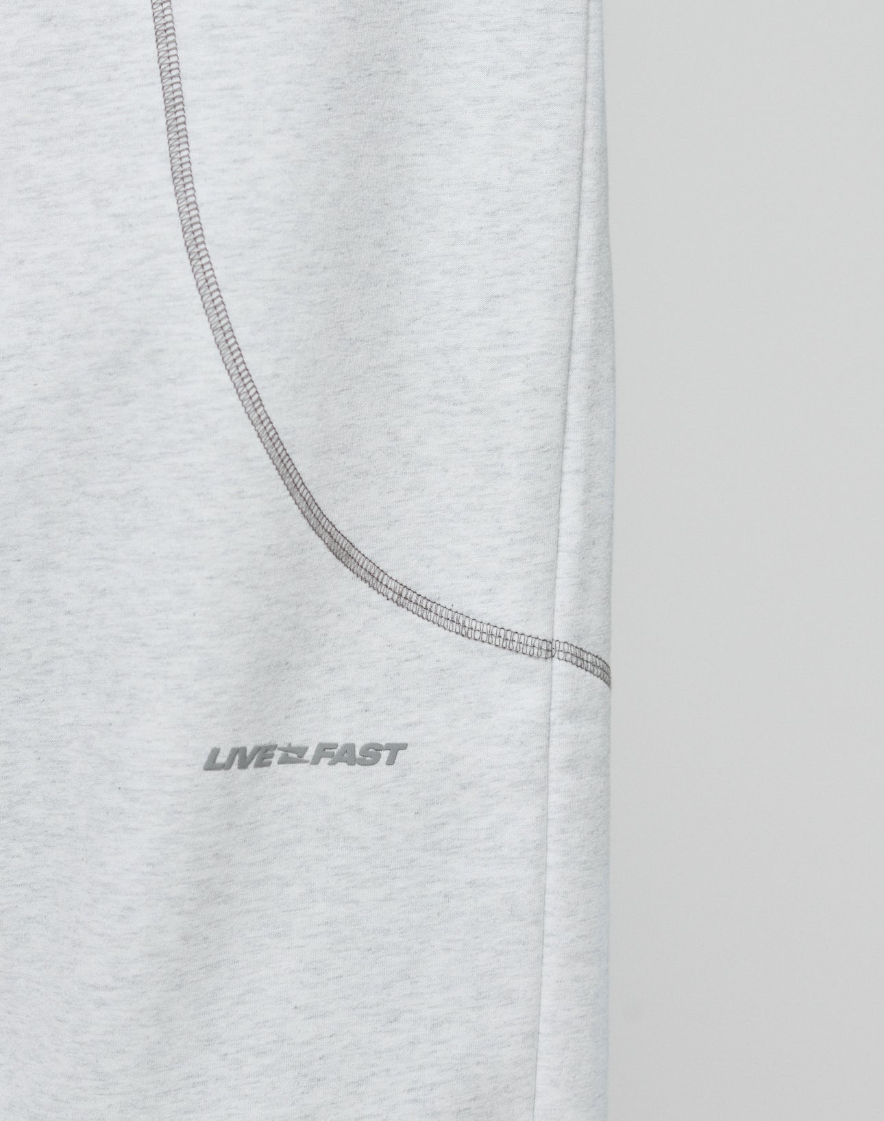 Close-up of the LFDY Active Fleece Jogger by LIVE FAST DIE YOUNG, featuring light gray fabric, a curved seam, and a subtle LIVE FAST logo in small gray uppercase letters. The background is plain and light-colored.