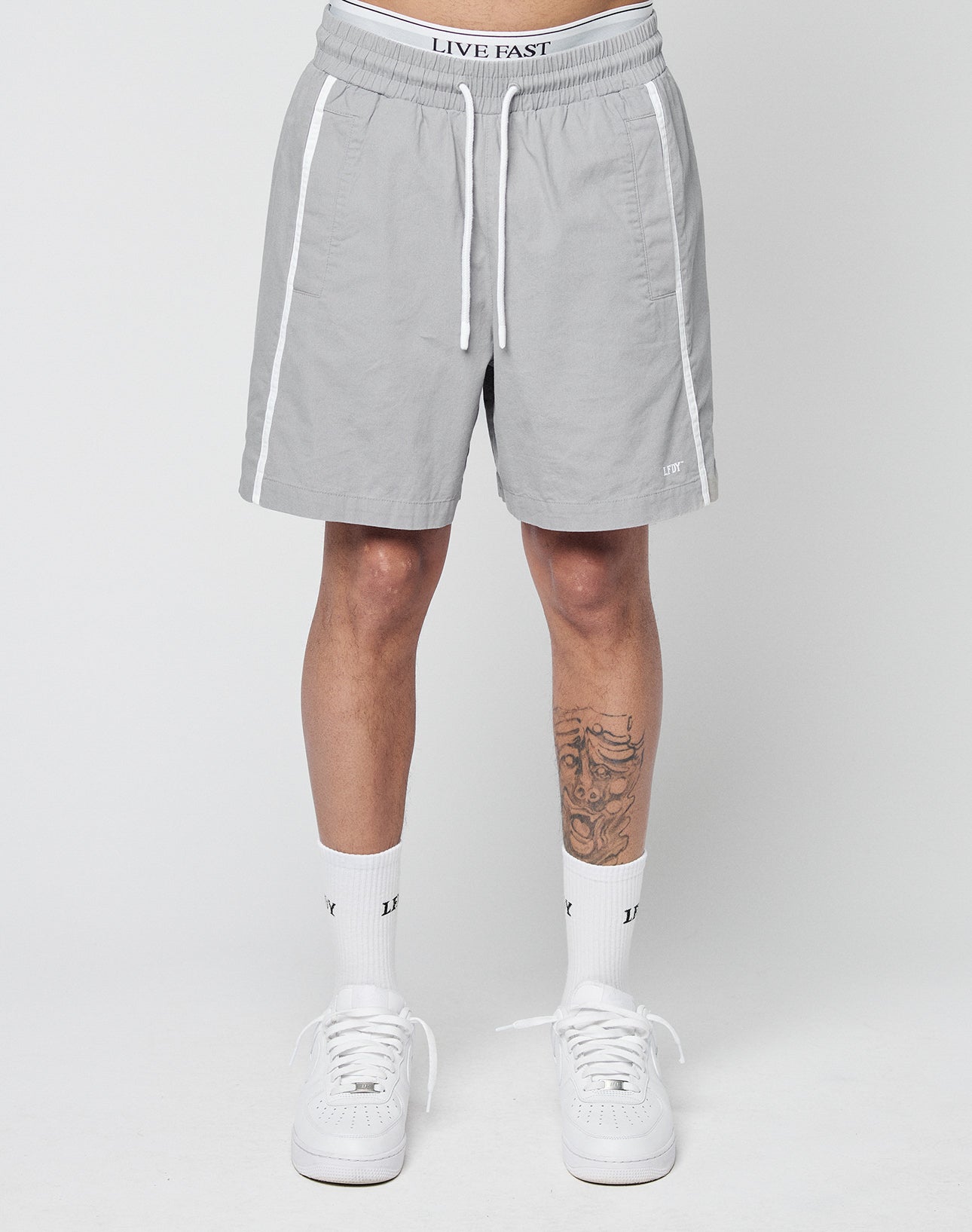 A person wearing LIVE FAST DIE YOUNG Basic Cotton Shorts in light gray with an elastic waistband featuring a LIVE FAST logo, white trim, white socks, and white sneakers stands against a plain background; a tattoo is visible on their left leg.