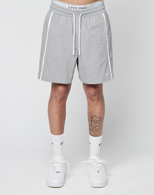 A person wearing LIVE FAST DIE YOUNG Basic Cotton Shorts in light gray with an elastic waistband featuring a LIVE FAST logo, white trim, white socks, and white sneakers stands against a plain background; a tattoo is visible on their left leg.