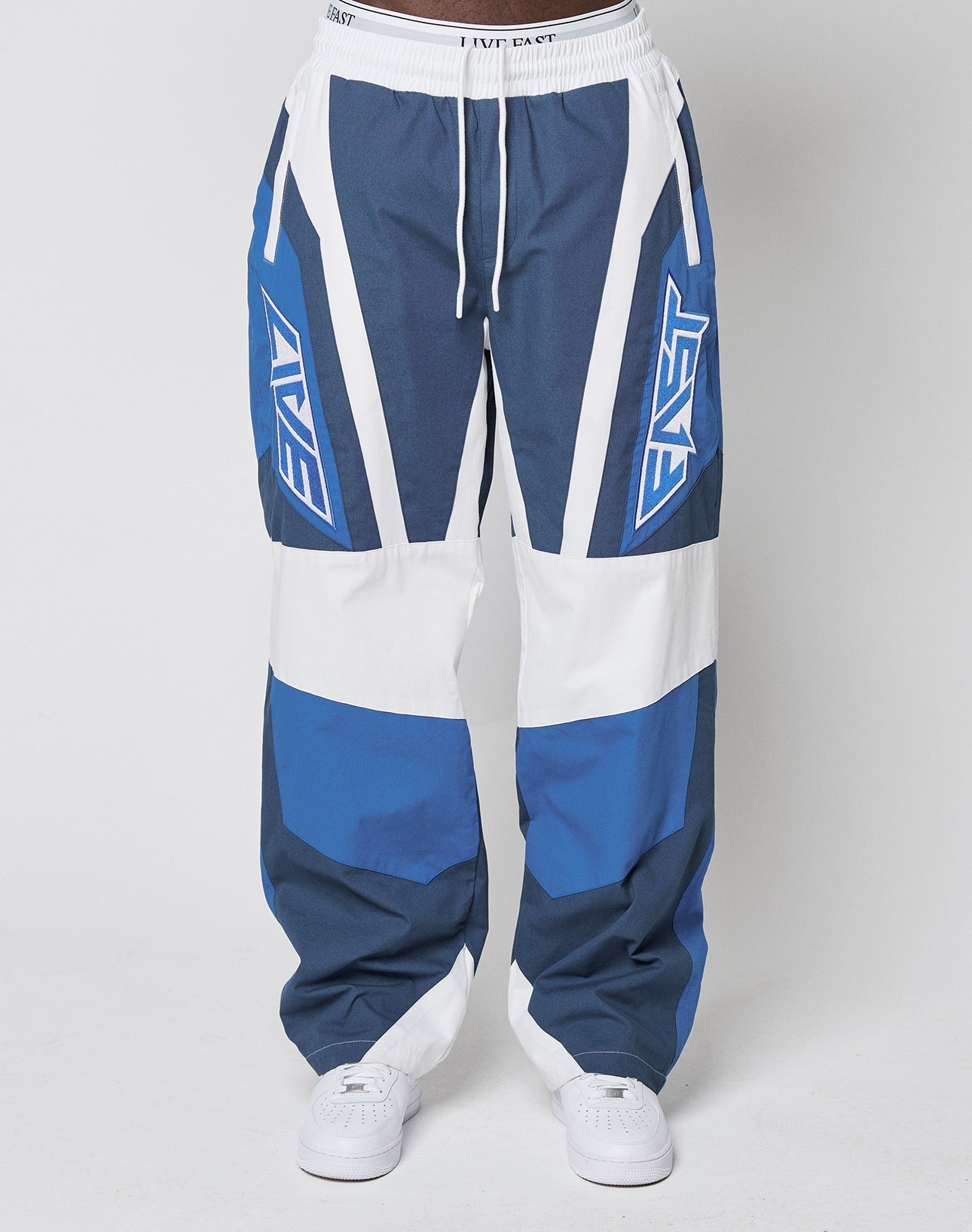 A person wears LIVE FAST DIE YOUNG LFDY Cross Pants—loose, color-blocked blue and white track pants with bold geometric patterns and EAST on the sides—paired with white sneakers for an oversized fit.