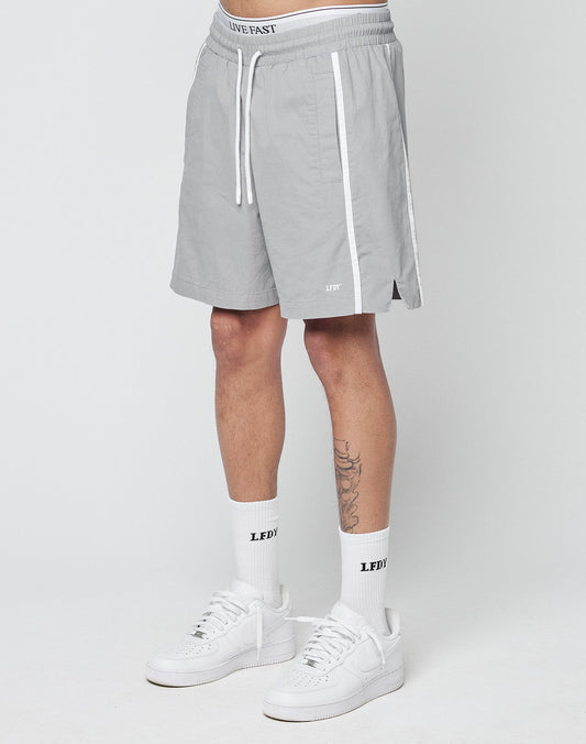 A person wearing LIVE FAST DIE YOUNG Basic Cotton Shorts in light gray with white stripes, elastic waistband featuring the LIVE FAST text, paired with LFDY logo socks and white sneakers, stands against a plain white background.