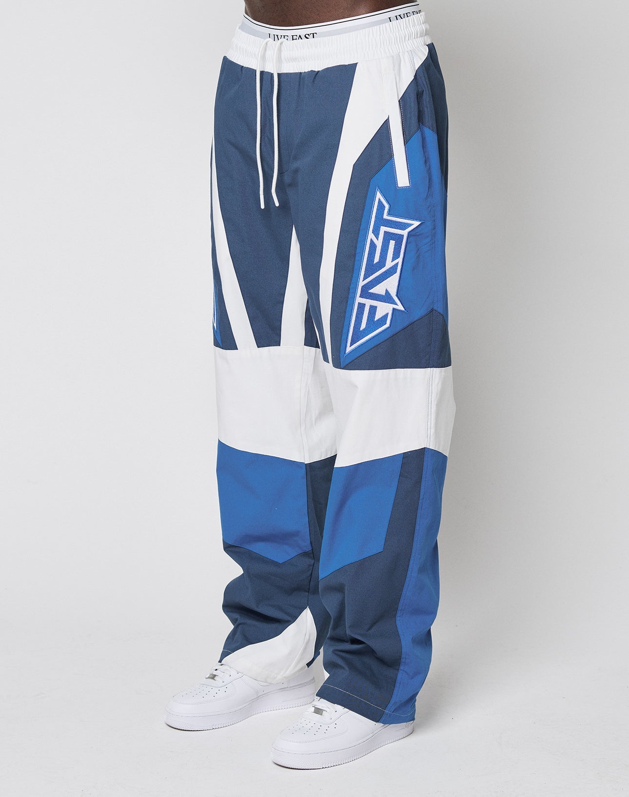 A person wears oversize LFDY Cross Pants by LIVE FAST DIE YOUNG, featuring blue, white, and navy color blocking with a bold “EAST” graphic on one leg and an elastic waistband, styled with white sneakers against a white background.