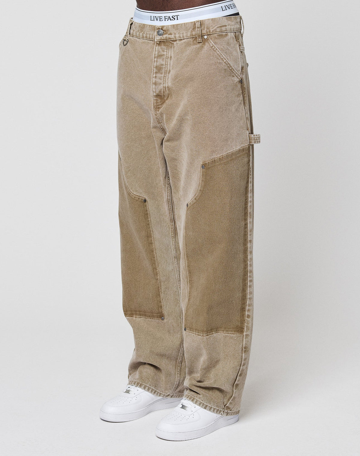 Someone stands against a plain light background wearing LIVE FAST DIE YOUNG Faded Carpenter Pants in a baggy fit, light brown color with visible seams, styled with white sneakers and a white LIVE FAST waistband. | model