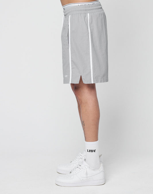 A person stands sideways wearing LIVE FAST DIE YOUNG Basic Cotton Shorts in light gray with white stripes and elastic waistband, white LFDY socks, and white sneakers against a plain background.