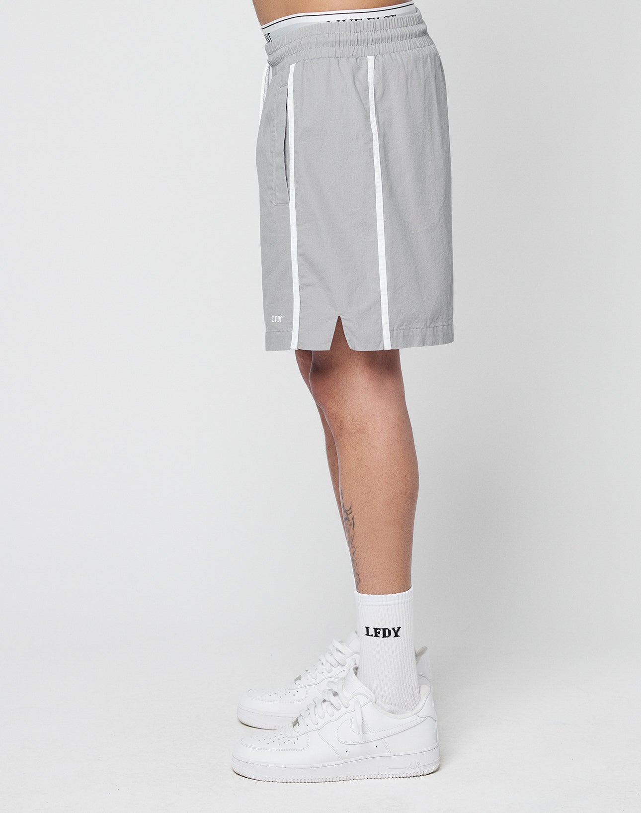 A person stands sideways wearing LIVE FAST DIE YOUNG Basic Cotton Shorts in light gray with white stripes and elastic waistband, white LFDY socks, and white sneakers against a plain background.