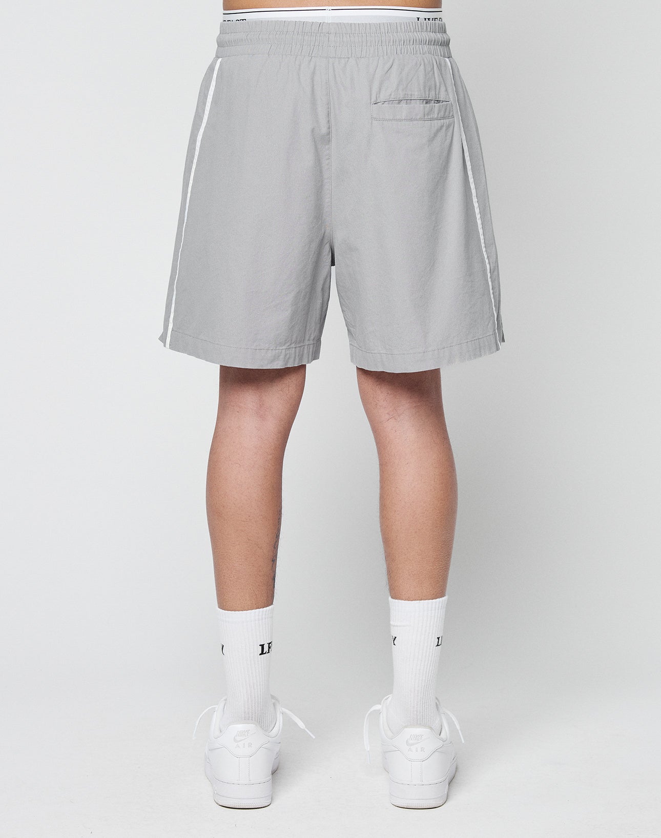 A person stands facing away in LIVE FAST DIE YOUNG Basic Cotton Shorts—light gray regular fit with white side stripes and elastic waistband—paired with white crew socks and sneakers, set against a plain light background.