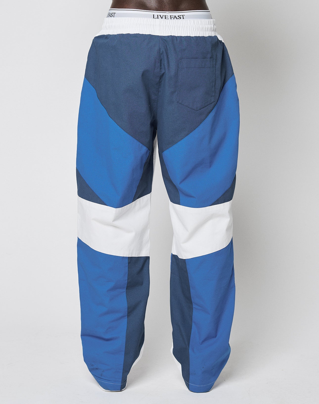 A person seen from the back wears LIVE FAST DIE YOUNGs LFDY Cross Pants—blue and white color-blocked with a geometric pattern, an Elastikbund waistband reading LIVE FAST, one right-side pocket, and crafted from soft Baumwolle.