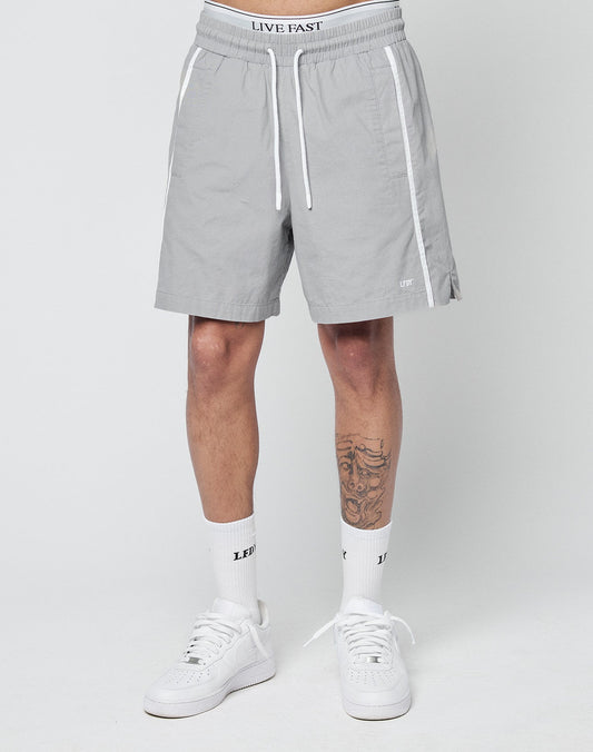 A person is shown from the waist down wearing LIVE FAST DIE YOUNG Basic Cotton Shorts in light gray, white sneakers, crew socks with LE and FT logos, and has a tattoo on one leg.
