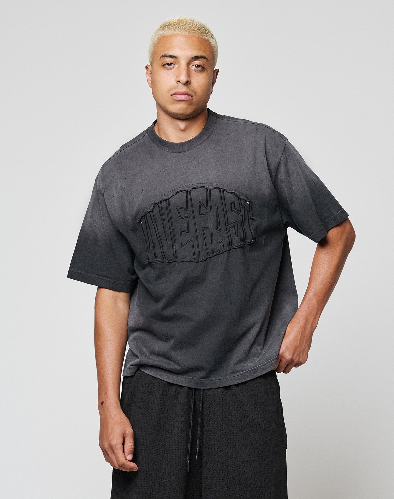 A young man with short bleached blond hair wears a loose, faded black LFDY Emblem Patch Tee by LIVE FAST DIE YOUNG and black shorts, posing with one hand in his pocket against a plain light background.
