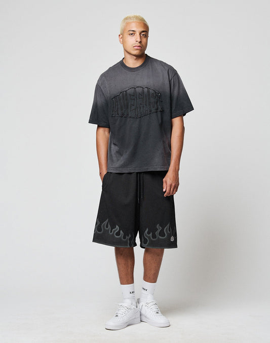 A young man with short blond hair wears a loose black T-shirt, LIVE FAST DIE YOUNG Burning Mesh Shorts with Elastikbund and Flammenprint at the hem, white crew socks, and sneakers, standing against a plain gray background.