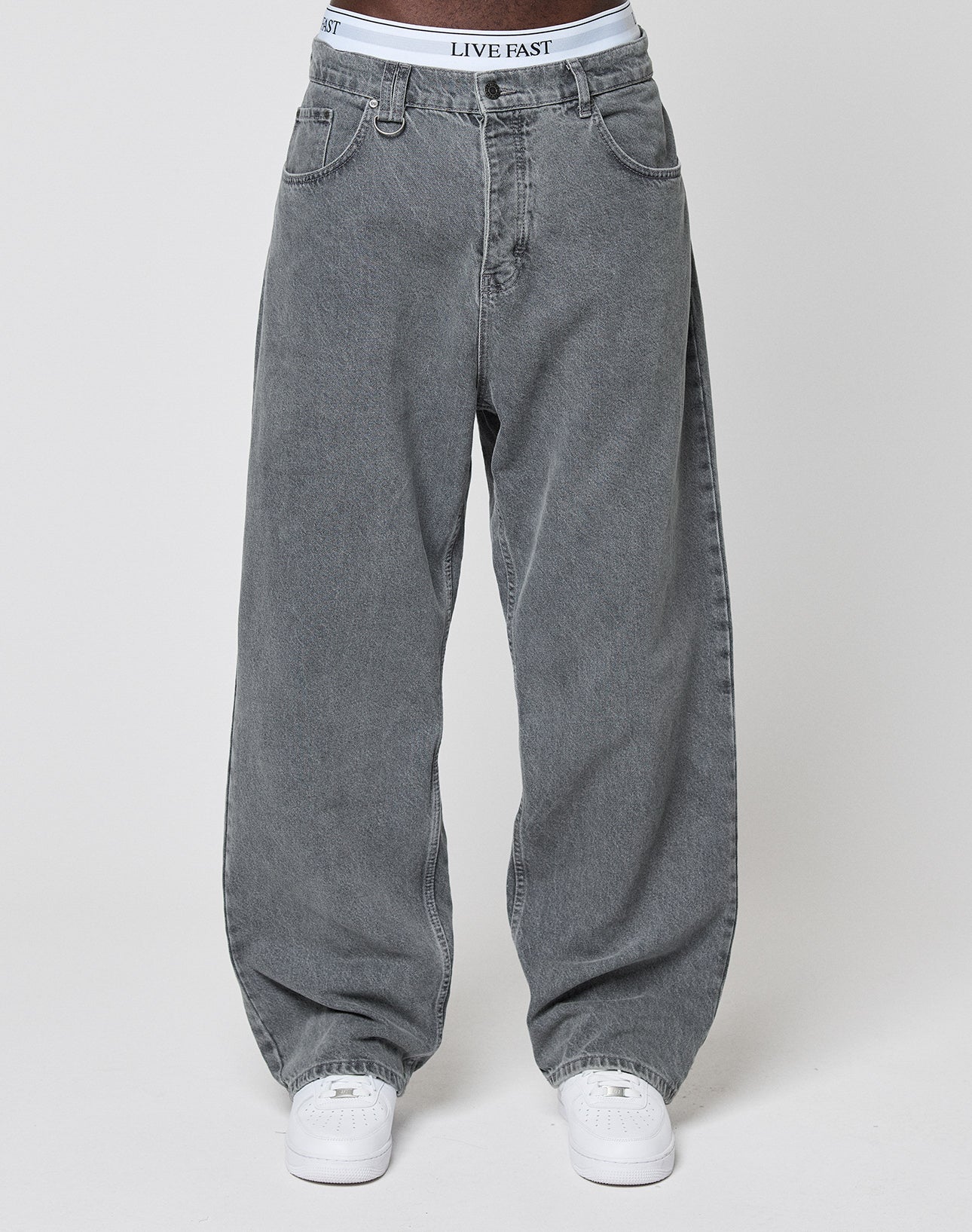 Wearing LFDY Baggy Denim by LIVE FAST DIE YOUNG—loose, light gray jeans with straight legs and visible front pockets, a LIVE FAST waistband, styled with white sneakers against a plain, light background.