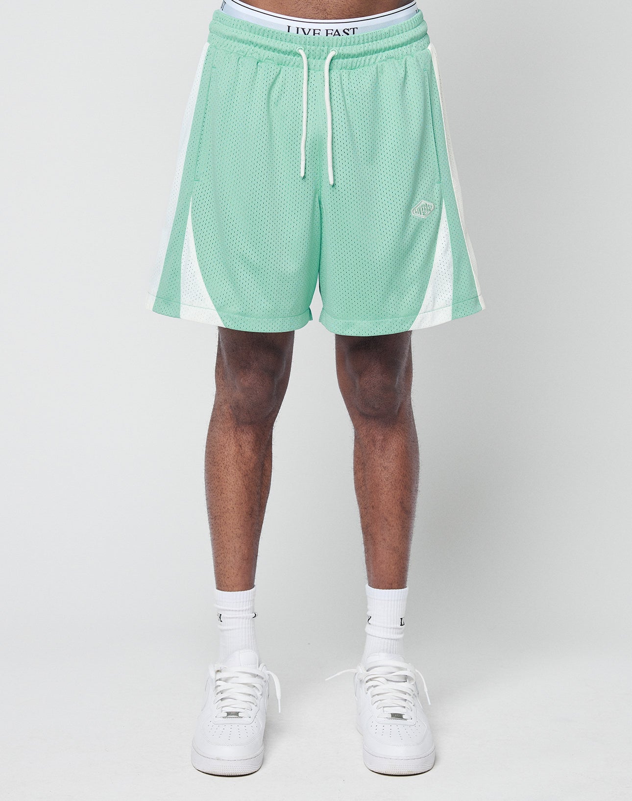 A person wears LIVE FAST DIE YOUNG Striped Mesh Shorts in mint green with logo patch and white side panels, styled in a regular fit with white crew socks and sneakers, standing against a plain light gray background.