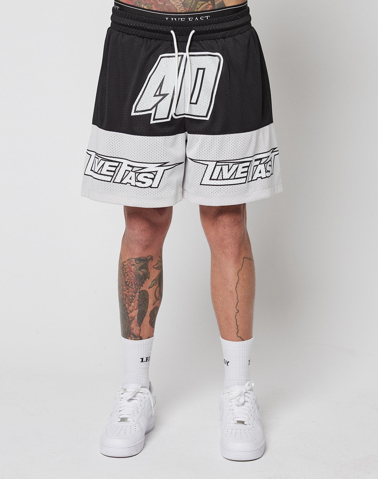 A person wears LFDY Racing Mesh Shorts by LIVE FAST DIE YOUNG, featuring a black-and-white design with “40” and “LIVE FAST” prints, a Regular Fit, and elastic waistband. Paired with white socks, white sneakers, and visible leg tattoos.