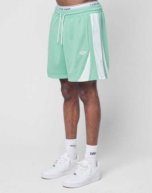 A person wearing LIVE FAST DIE YOUNG Striped Mesh Shorts in mint green with white stripes and logo patch, paired with white LFDY socks and white Nike sneakers, stands against a light background. The shorts feature a regular fit and breathable polyester. | model