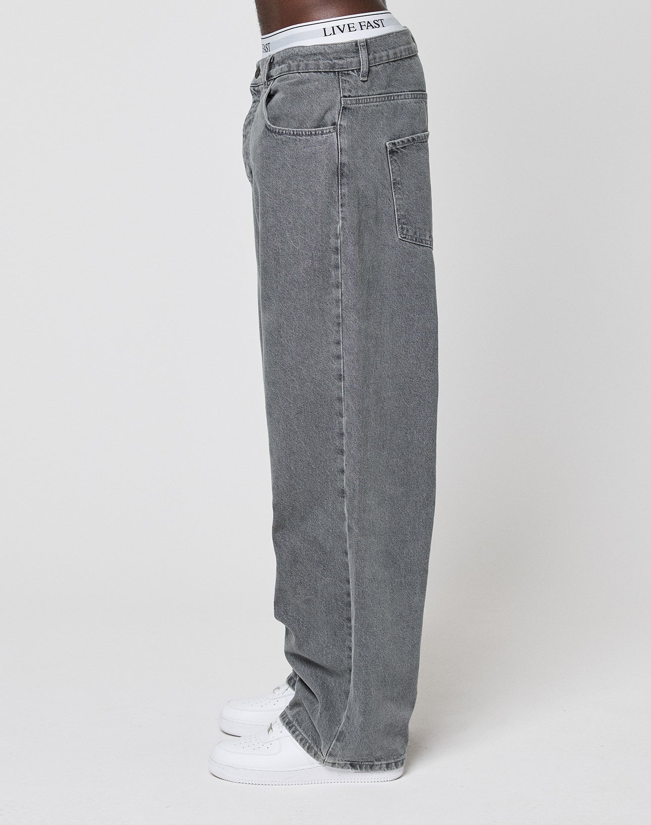 A person stands sideways in loose-fitting, high-waisted LFDY Baggy Denim jeans by LIVE FAST DIE YOUNG, paired with white sneakers and LIVE FAST underwear, against a plain white background.
