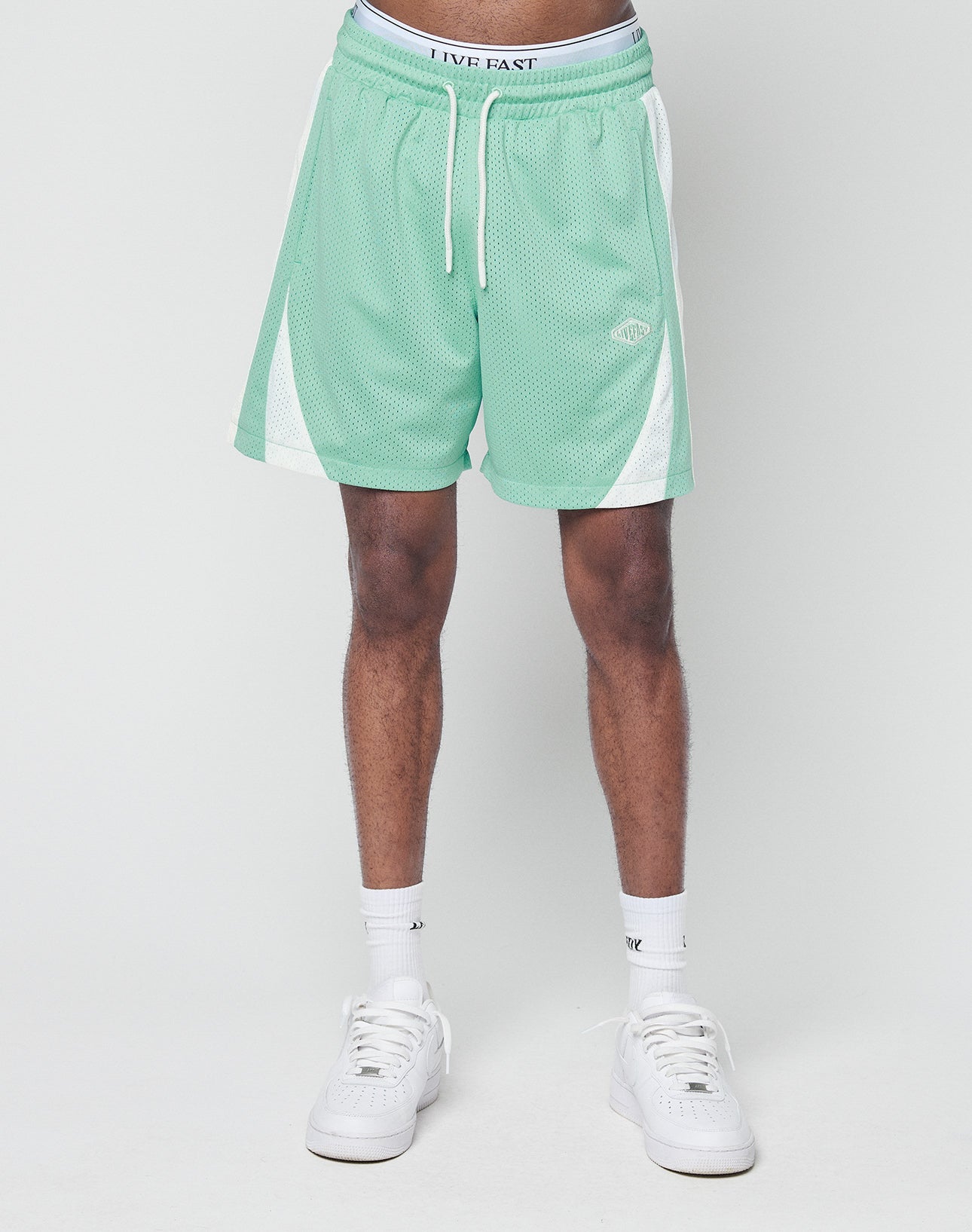 A person sports LIVE FAST DIE YOUNG Striped Mesh Shorts in mint green with white side panels and a subtle logo patch, styled in a regular fit with white socks and sneakers, standing against a plain light gray background. Only the lower half is visible.