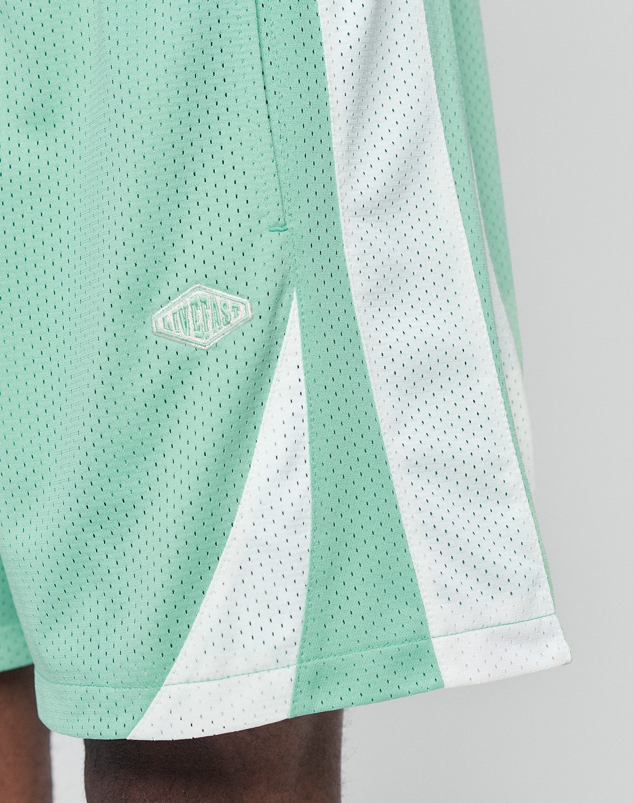 A close-up of someone wearing LIVE FAST DIE YOUNG Striped Mesh Shorts in mint green and white with a logo patch on the left, partially showing their leg and the shorts regular fit.