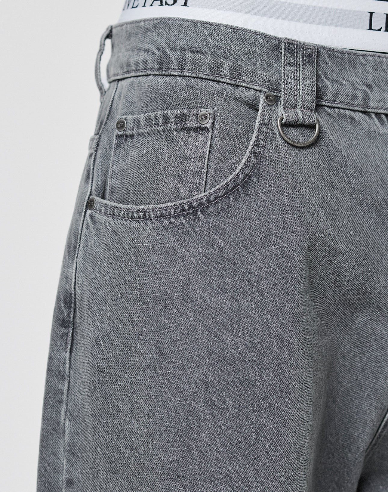 Close-up of a person wearing LFDY Baggy Denim jeans by LIVE FAST DIE YOUNG in light gray with baggy fit, straight leg, small front pocket, belt loops, silver D-ring, and white underwear waistband with black text peeking above.