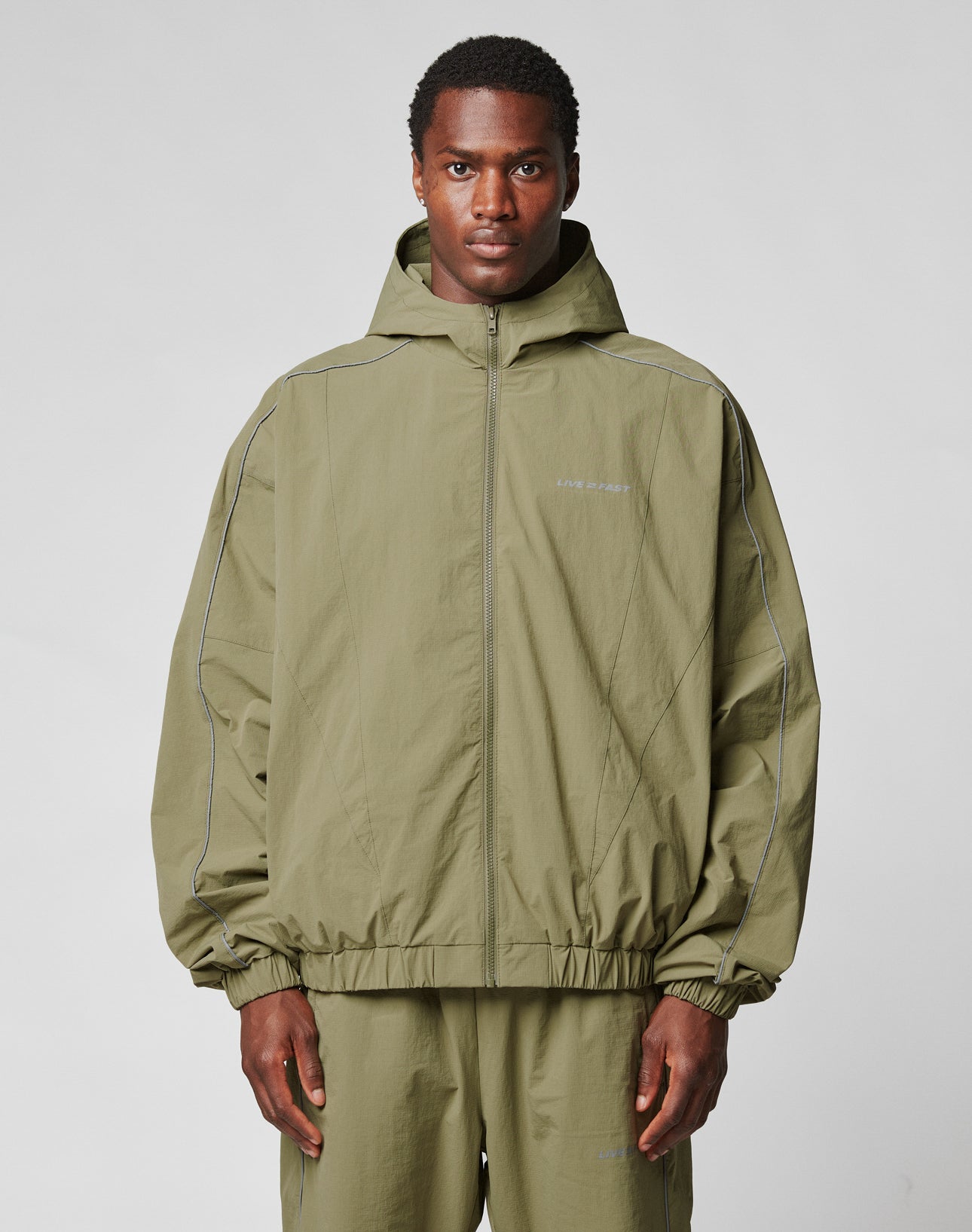 A man wears the LIVE FAST DIE YOUNG Live Fast Trackjacket in olive green nylon with reflective piping, a front zipper, and matching pants, standing against a plain light gray background.