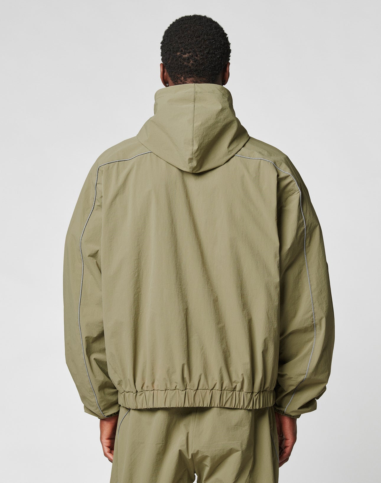 A person is seen from behind wearing the LIVE FAST DIE YOUNG Live Fast Trackjacket in light olive green nylon with elastic waist, visible stitched seams, and reflective pipings, set against a plain white background.